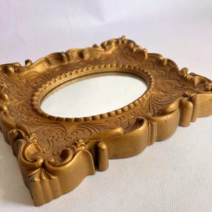Puede incluir: Un espejo ovalado dorado con un marco ornamentado y detallado. El marco presenta patrones florales en relieve y elementos decorativos. El espejo se encuentra sobre un fondo blanco, mostrando su est&eacute;tica vintage.