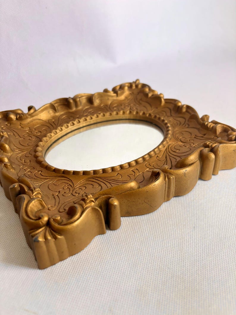 Puede incluir: Un espejo ovalado dorado con un marco ornamentado y detallado. El marco presenta patrones florales en relieve y elementos decorativos. El espejo se encuentra sobre un fondo blanco, mostrando su est&eacute;tica vintage.