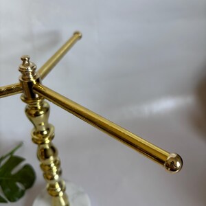 May include: A gold-colored jewelry display stand with a white and gray marble base. The stand has a decorative, ornate post and three horizontal bars for hanging necklaces or bracelets. The ends of the bars have small, round gold accents.