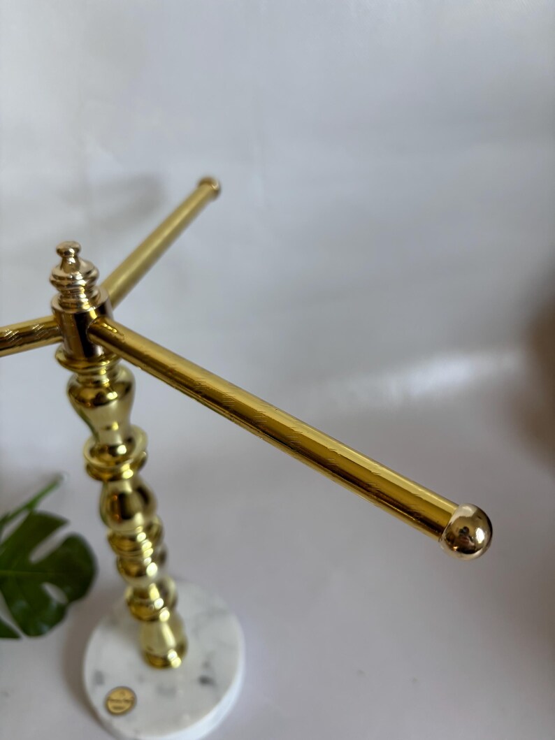 May include: A gold-colored jewelry display stand with a white and gray marble base. The stand has a decorative, ornate post and three horizontal bars for hanging necklaces or bracelets. The ends of the bars have small, round gold accents.