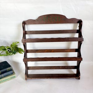 Op de afbeelding: Een donkerbruin, vintage houten wandrek met vier horizontale latten. De bovenkant van het rek heeft een decoratief, gebogen ontwerp met vervaagde groene tekst. Het rek is ontworpen om items op te bergen.
