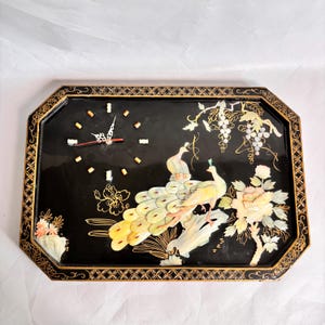 May include: A rectangular black wall clock with an octagonal frame featuring gold geometric patterns. The clock face displays two peacocks and a floral arrangement crafted from iridescent shell inlays, alongside gold-toned hour markers.