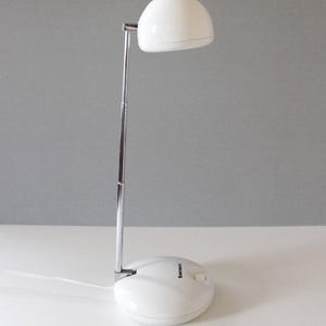 May include: A white desk lamp with a rounded shade and a chrome-colored adjustable arm. The base is oval-shaped with a power button. The lamp is plugged into a white surface.