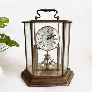 May include: A vintage brass-toned quartz clock with a handle. The clock has a white face with black Roman numerals and a pendulum with three silver spheres. The clock is encased in glass panels with a decorative etched design.