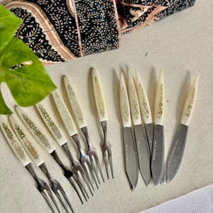 Puede incluir: Un conjunto de cubiertos antiguos, que incluye tenedores y cuchillos, con mangos de color marfil adornados con diseños florales dorados. Las hojas de acero inoxidable de los cuchillos son visibles. Un tejido estampado en tonos azul oscuro, rosa y beige está en el fondo.