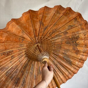 Puede incluir: Un parasol de papel naranja abierto con un mango de madera. La superficie del parasol presenta un patr&oacute;n oscuro y detallado. Las varillas son de madera fina y clara. El mango es de color marr&oacute;n claro.