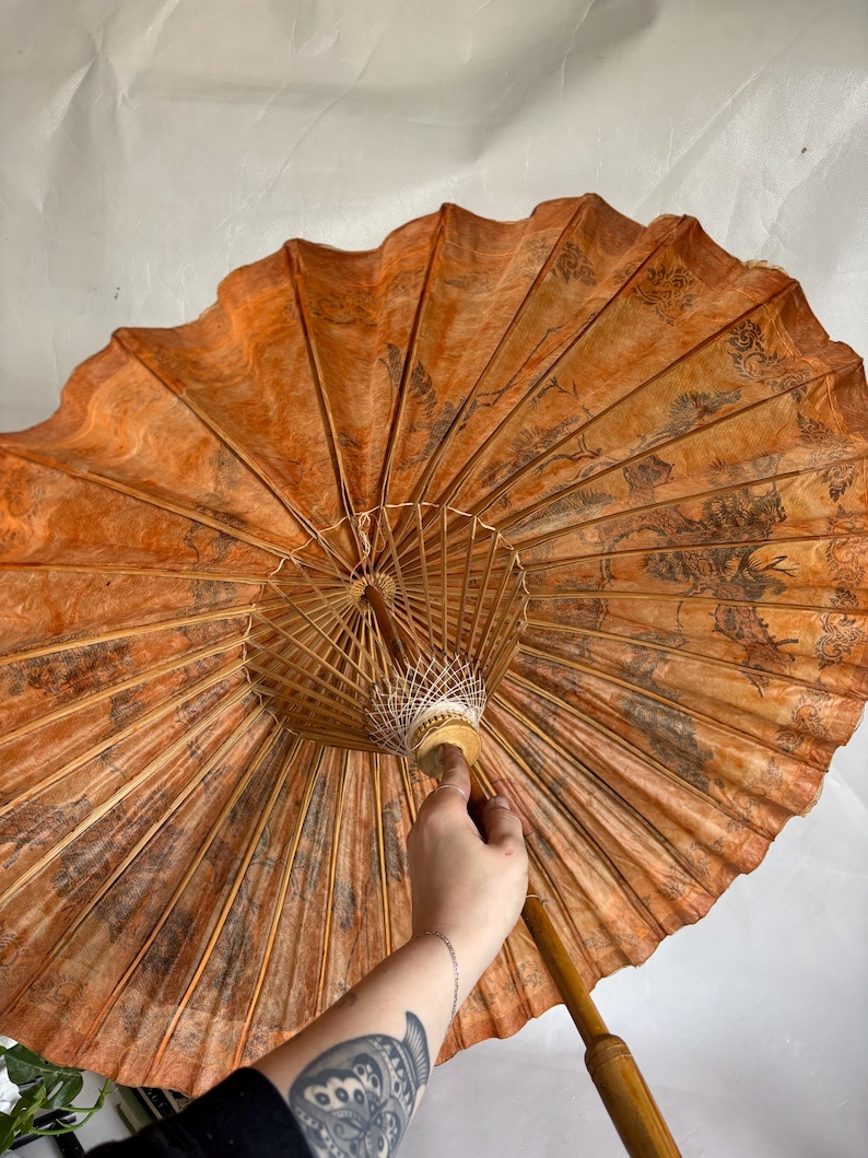 Puede incluir: Un parasol de papel naranja abierto con un mango de madera. La superficie del parasol presenta un patr&oacute;n oscuro y detallado. Las varillas son de madera fina y clara. El mango es de color marr&oacute;n claro.