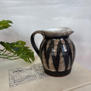 Könnte beinhalten: Ein kleiner Keramikkrug mit schwarzem Henkel und Ausguss. Der Körper weist braune und schwarze vertikale Zickzackmuster auf. Der Innenraum ist weiß und der Boden ist schwarz. Der Krug wird wahrscheinlich zum Servieren von Flüssigkeiten verwendet.