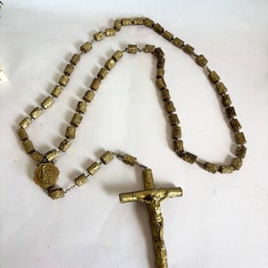 May include: A gold-colored rosary with a crucifix. The rosary beads are rectangular and connected by small metal links. The crucifix features a detailed depiction of Jesus. The rosary is displayed on a white surface.