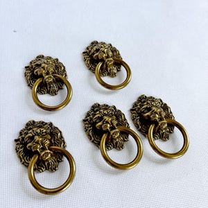 May include: Five antique brass lion head drawer pulls with circular handles. The lion heads have detailed manes and are mounted on a white surface. These decorative pulls are ideal for adding a vintage touch to furniture.