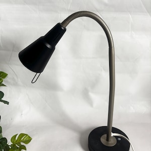 Puede incluir: Una lámpara de escritorio negra con un cuello de cisne metálico flexible y una base redonda. La lámpara tiene una pantalla cónica y un pequeño asa curvada. La base es negra y el cuello de cisne es de color metálico oscuro.