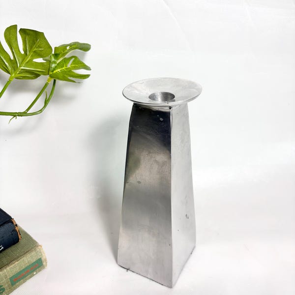 May include: A silver-colored metal candle holder with a tall, four-sided tapered base and a wide, circular top opening. The object features a smooth, reflective finish and a geometric, pyramid-like structure that stands upright on a flat surface.