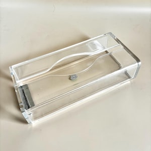 Puede incluir: Un porta-cajas de pañuelos de acrílico transparente. La caja rectangular tiene una abertura curva en la parte superior para facilitar el acceso a los pañuelos. Una pequeña pieza de metal plateado está unida al lateral. El soporte está diseñado para una caja de pañuelos estándar.