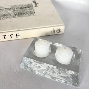 Puede incluir: Un portavelas rectangular transparente con dos velas blancas. El soporte está hecho de un material texturizado, posiblemente de vidrio. Un libro con el título "ETTE" está en el fondo.