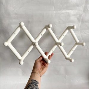 May include: A white, expandable wall-mounted coat rack. The rack is made of white plastic and features a series of diamond-shaped sections with rounded knobs at the ends. The rack is held by a hand with a tattoo on the arm.