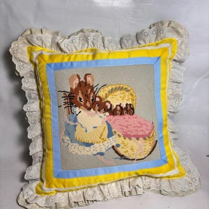 May include: A square decorative pillow featuring a needlepoint scene of a brown mouse tending to smaller mice in a yellow cradle. The central panel is framed by blue and yellow fabric borders and finished with a white eyelet lace ruffled edge.