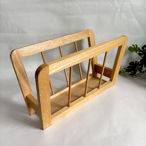 Puede incluir: Un revistero de madera marrón claro con forma rectangular. El revistero tiene un diseño de listones en los laterales y una base sólida. El marco es de madera de color claro y el diseño general es sencillo y moderno.