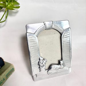 May include: A silver-toned metal picture frame shaped like an arched window with open shutters. The frame features a raised relief design at the base, depicting a potted plant on the left and a cat resting on the right side of the window sill.