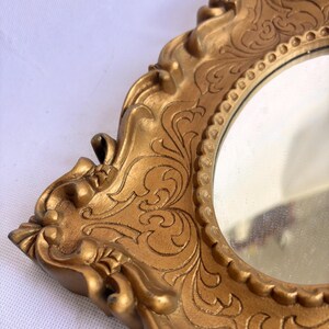 Puede incluir: Primer plano de un espejo dorado y ornamentado con una superficie reflectante redonda. El marco presenta detalles de volutas y un dise&ntilde;o decorativo vintage. El espejo es probablemente para fines decorativos.