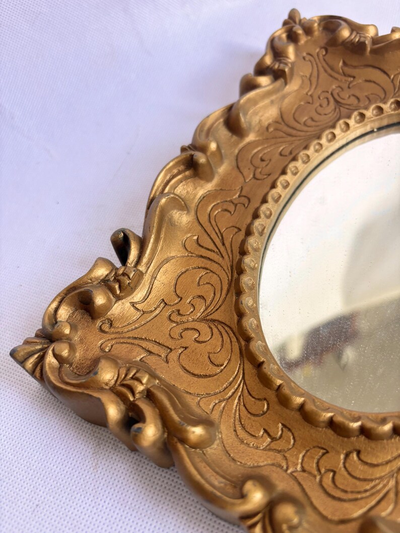 Puede incluir: Primer plano de un espejo dorado y ornamentado con una superficie reflectante redonda. El marco presenta detalles de volutas y un dise&ntilde;o decorativo vintage. El espejo es probablemente para fines decorativos.