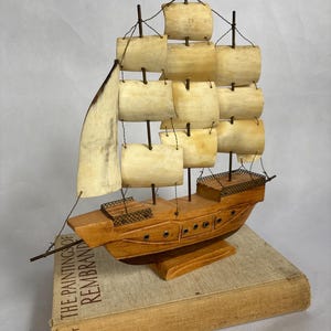 May include: A wooden model ship with multiple sails and rigging, resting on a book titled "The Paintings of Rembrandt." The ship is crafted from light brown wood, with off-white sails and dark brown rigging. The book has a beige cover.