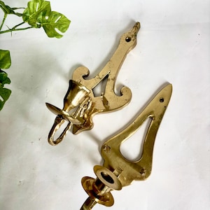 Pode incluir: Dois castiçais de latão vintage. Os castiçais têm um design ornamentado com uma forma curva e uma base circular para segurar a vela. O latão tem um tom dourado quente. O fundo é uma superfície branca.