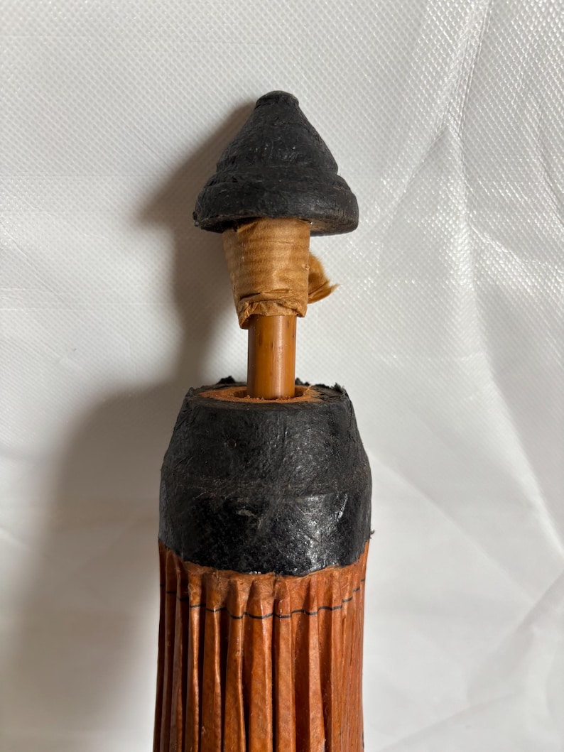 Puede incluir: Primer plano de un paraguas vintage. El paraguas presenta una parte superior negra en forma de cono y un mango de madera marr&oacute;n. El cuerpo del paraguas est&aacute; hecho de material plisado de color marr&oacute;n rojizo, con detalles en negro. El paraguas es un dise&ntilde;o cl&aacute;sico.