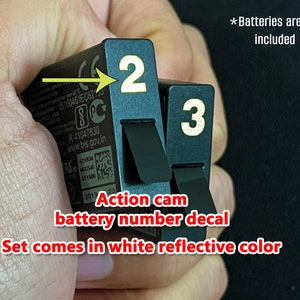 May include: Close-up of two black battery number decals with white reflective numbers. The decals are labeled "2" and "3". The text "Action cam battery number decal" and "Set comes in white reflective color" is written in red below the decals.