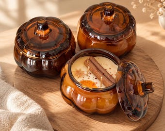 May include: Three pumpkin-shaped amber glass jars, two with lids on and one open. The open jar contains cream-colored wax with two wicks, a cinnamon stick, and a dried orange slice. The glass has a ribbed texture and a small stem-like handle on top.