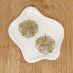 May include: Two translucent, glittery, shell-shaped resin charms with gold-toned metal jump rings. One charm features the name PADME in gold lettering, and the other features the name ROCKET. Both are decorated with small gold and white starfish and shell accents.