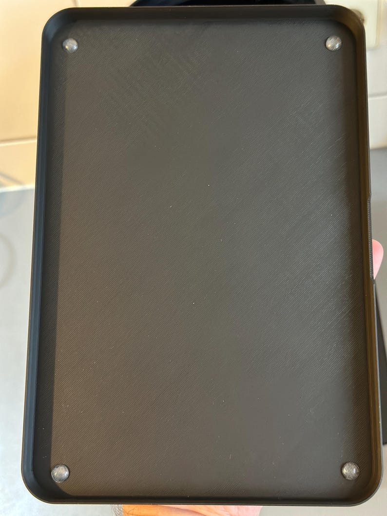 May include: A rectangular black tray with a textured surface and rounded corners. Four silver-colored rivets are visible, one in each corner. The tray appears to be made of a durable material.
