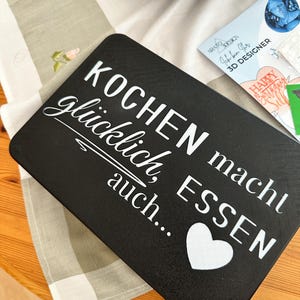 May include: A black rectangular cutting board with white text that reads "KOCHEN macht gl&uuml;cklich, auch... ESSEN" and a white heart. The board is on a wooden table with a floral patterned tablecloth.