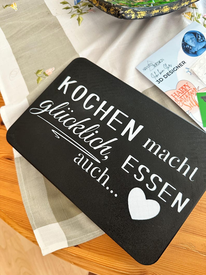 May include: A black rectangular cutting board with white text that reads "KOCHEN macht gl&uuml;cklich, auch... ESSEN" and a white heart. The board is on a wooden table with a floral patterned tablecloth.