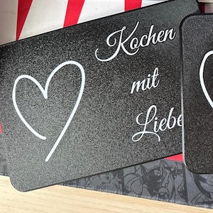 May include: Black rectangular kitchen cutting board with a white heart outline and the German text "Kochen mit Liebe" (Cooking with Love). The board has a textured surface.