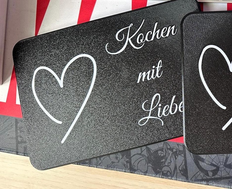 May include: Black rectangular kitchen cutting board with a white heart outline and the German text "Kochen mit Liebe" (Cooking with Love). The board has a textured surface.