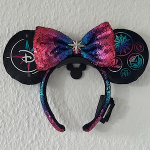 Könnte beinhalten: Schwarze Mickey Mouse Ohren mit einer paillettenbesetzten Schleife in Rosa, Blau und Lila. Die Ohren zeigen Disney-Motive, darunter ein Kompass, ein Schneeflocke und das Avengers-Logo. Ein silberner Stern ist auf der Schleife zentriert.