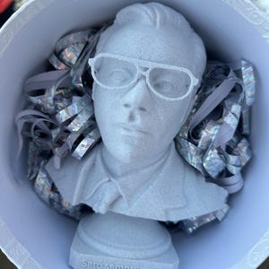 Könnte beinhalten: Eine weiße Büste einer Person mit Brille, die in einer weißen Schüssel mit silbernen und weißen Konfetti sitzt. Die Büste trägt die Worte "Saro sempre con te" auf dem Sockel.