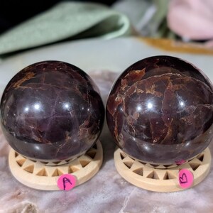 May include: Two polished, dark red-brown gemstone spheres, each resting on a small, light-colored wooden stand. The spheres have a glossy finish with internal patterns. Pink stickers labeled "A" and "B" are attached to the stands. The background is blurred.