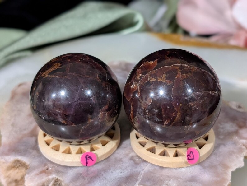 May include: Two polished, dark red-brown gemstone spheres, each resting on a small, light-colored wooden stand. The spheres have a glossy finish with internal patterns. Pink stickers labeled "A" and "B" are attached to the stands. The background is blurred.
