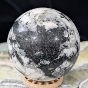 May include: A polished sphere with a marbled appearance, featuring a mix of black, white, and gray patterns. The orb is displayed on a small, round wooden stand. The sphere's surface has a glossy finish, reflecting light.