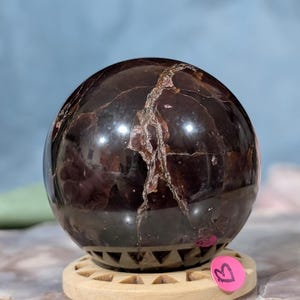 May include: A polished, dark red gemstone sphere with light brown veining. The sphere sits on a light brown wooden stand with a decorative geometric pattern. A pink sticker with the letter "B" is attached to the stand.