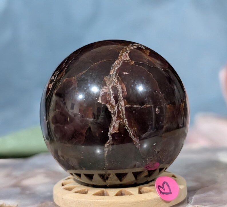 May include: A polished, dark red gemstone sphere with light brown veining. The sphere sits on a light brown wooden stand with a decorative geometric pattern. A pink sticker with the letter "B" is attached to the stand.