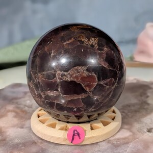 May include: A polished, dark red gemstone sphere with a textured surface, resting on a light wooden stand. The sphere has a mix of deep red and brown tones. A pink sticker with the letter "A" is on the stand.