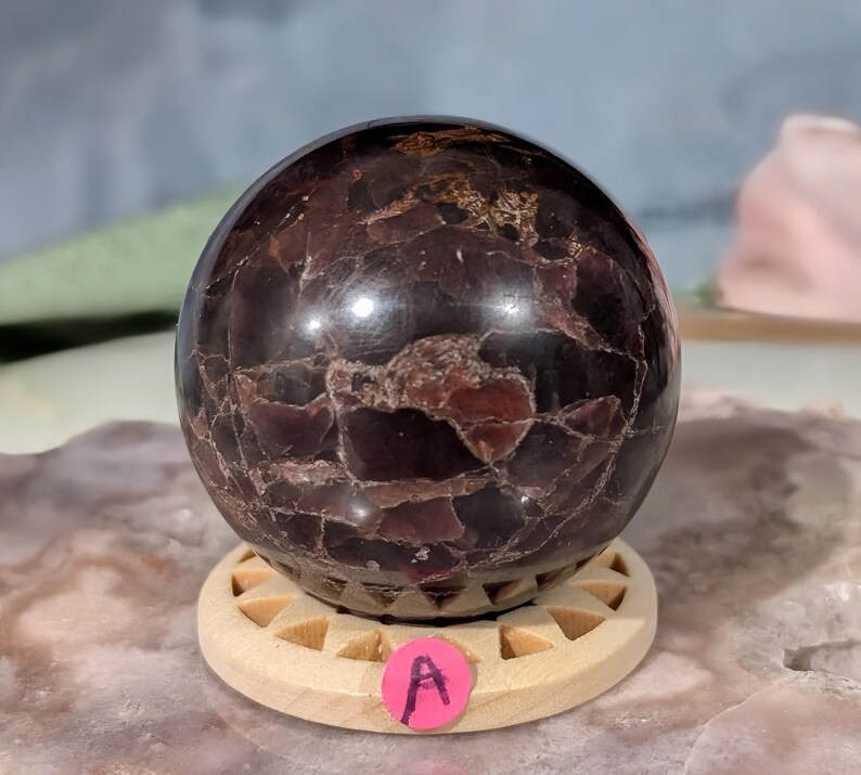 May include: A polished, dark red gemstone sphere with a textured surface, resting on a light wooden stand. The sphere has a mix of deep red and brown tones. A pink sticker with the letter "A" is on the stand.