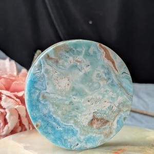 May include: A circular polished stone slab featuring a marbled pattern of vibrant turquoise, sky blue, and white hues with subtle tan and brown mineral inclusions throughout the surface, displaying a smooth, glossy texture and natural stone variations.