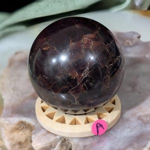 May include: A polished, dark red-brown gemstone sphere, resting on a light wood stand with triangular cutouts. The sphere has a reflective surface with natural patterns. A small pink sticker with the letter "A" is visible.