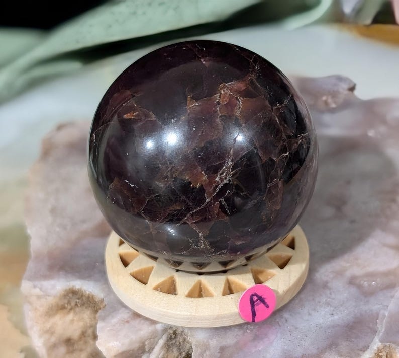 May include: A polished, dark red-brown gemstone sphere, resting on a light wood stand with triangular cutouts. The sphere has a reflective surface with natural patterns. A small pink sticker with the letter "A" is visible.