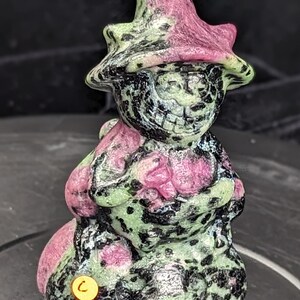 May include: A green and pink carved stone figurine of a gnome wearing a pointed hat. The gnome is holding a bag in its hand and has a whimsical expression on its face.