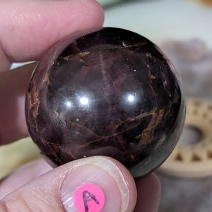 May include: A polished, dark purple sphere with brown veining. The gemstone is held in a hand, with a pink sticker labeled "A" visible. The sphere has a reflective surface, and the background is blurred.
