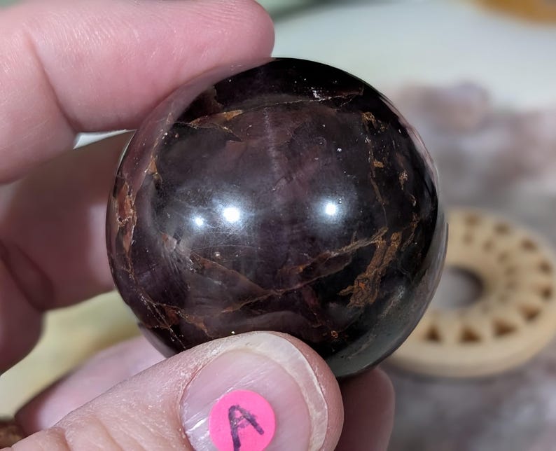 May include: A polished, dark purple sphere with brown veining. The gemstone is held in a hand, with a pink sticker labeled "A" visible. The sphere has a reflective surface, and the background is blurred.